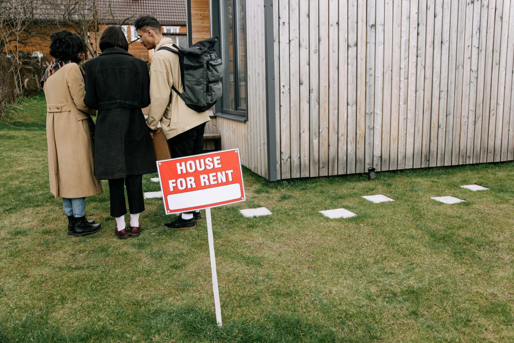 Three people seen from behind on a lawn with a building to the right. A House for Rent sign is in the foreground - Visit Afinestpm.com. 